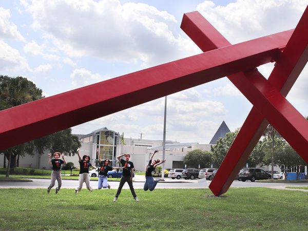 several people in mid jump under a large sculpture of intersecting red beams with a museum building in the distance