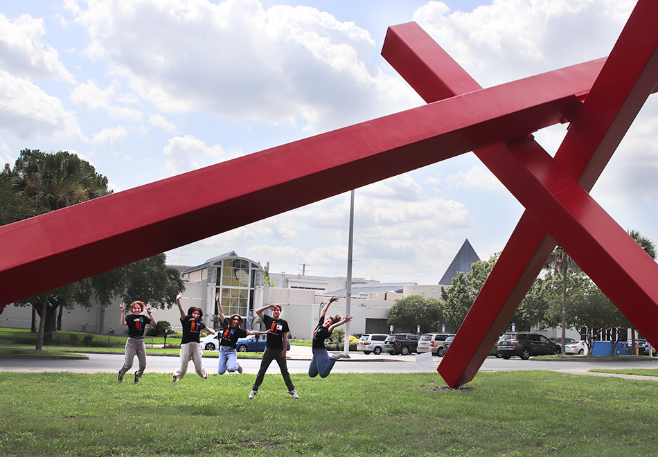 several people in mid jump under a large sculpture of intersecting red beams with a museum building in the distance