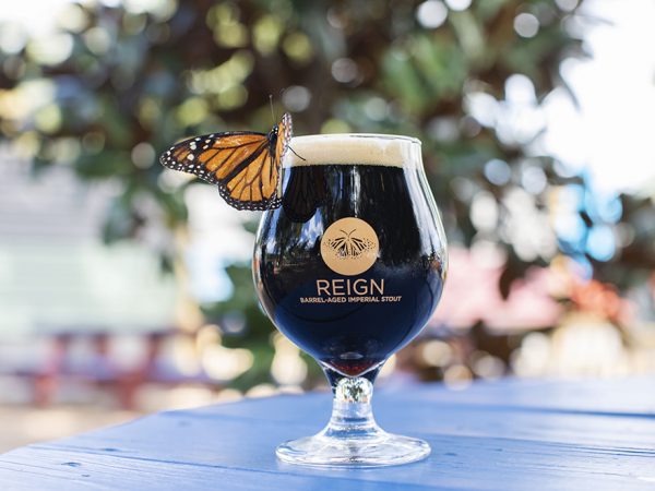 monarch butterfly on the lip of a glass of dark beer on a blue table. The glass says Reign Barrel-Aged Imperial Stout and has a picture of monarch on it