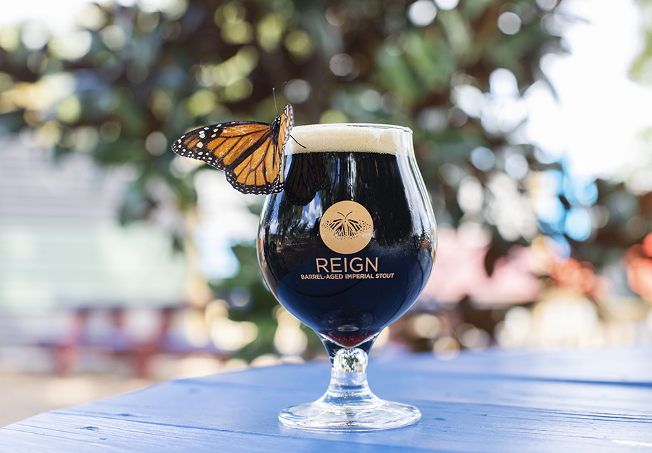 monarch butterfly on the lip of a glass of dark beer on a blue table. The glass says Reign Barrel-Aged Imperial Stout and has a picture of monarch on it