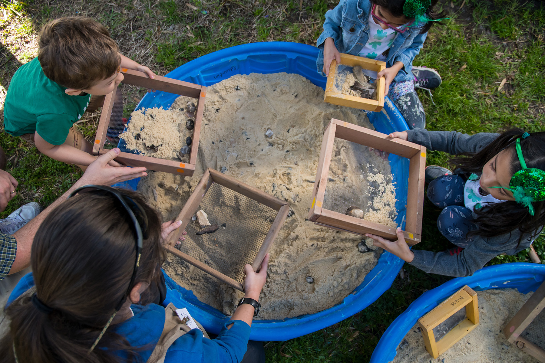 several young children kneel around a small plastic pool filled with sand and use screens to sift through the sand to find rock and fossils