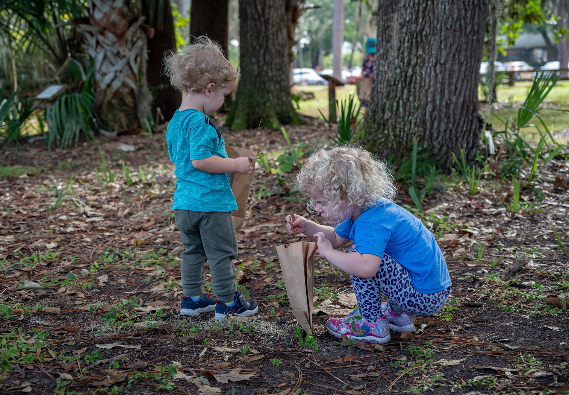 two young children outside holding brown paper bags, one child is kneeling down and placing sticks into the bag