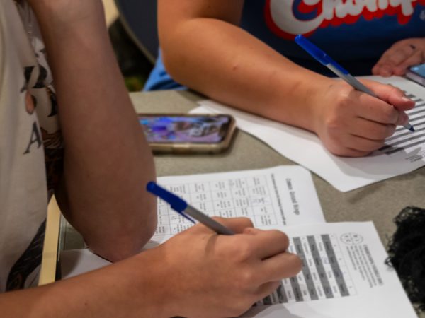 close up of a table and two people's hands with pens poised over papers printed with lists