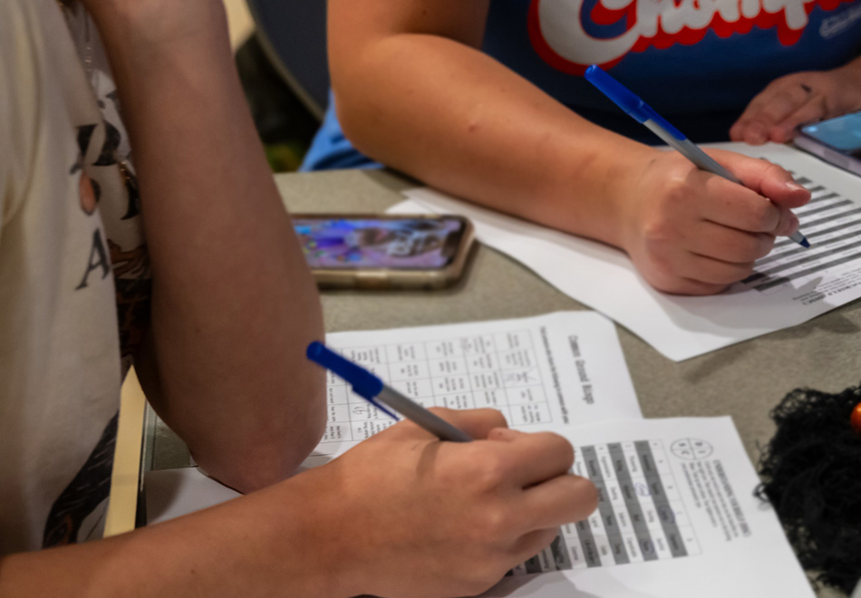 close up of a table and two people's hands with pens poised over papers printed with lists