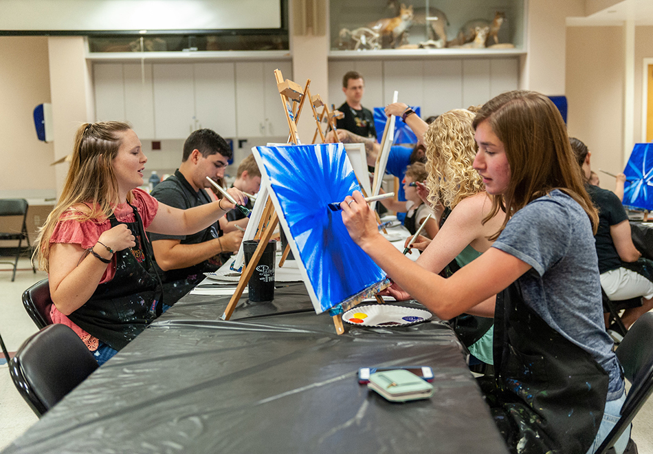 a group of people painting at long tables in a classroom setting