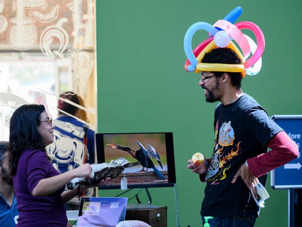 a person at a table is showing a large bird specimen to someone that is wearing an elaborate hat made out of balloons in a public space like a museum