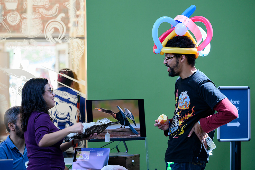a person at a table is showing a large bird specimen to someone that is wearing an elaborate hat made out of balloons in a public space like a museum