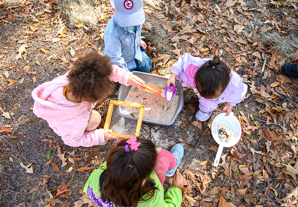 Children use a sifting screen, paint brushes, colanders, and shovels to find objects in sand during a Tot Trots activity.