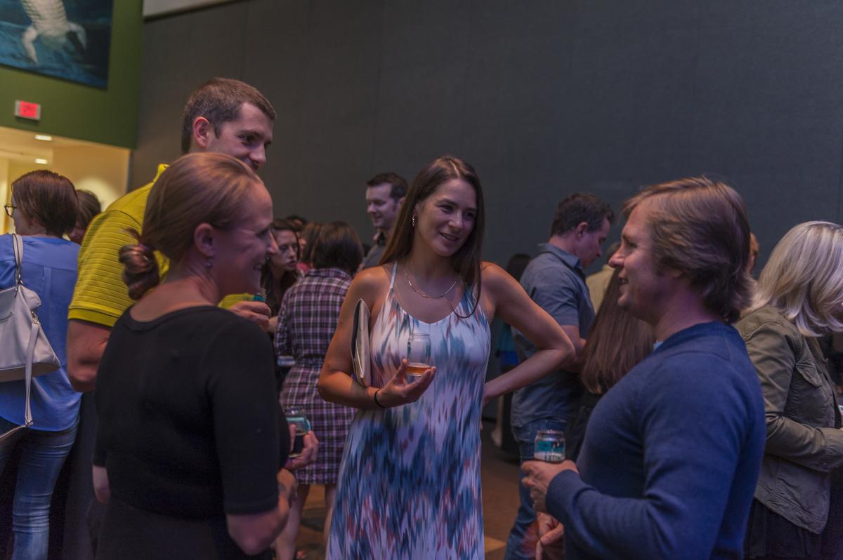 a group of people holding beer glasses are talking in a somewhat dark room in a public space as if at a party