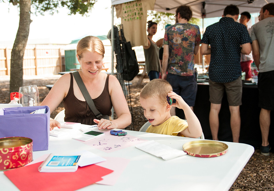 an adult and child sit at a table doing crafts with papers and stamps at an outdoors venue with people crowded around a tent in the background