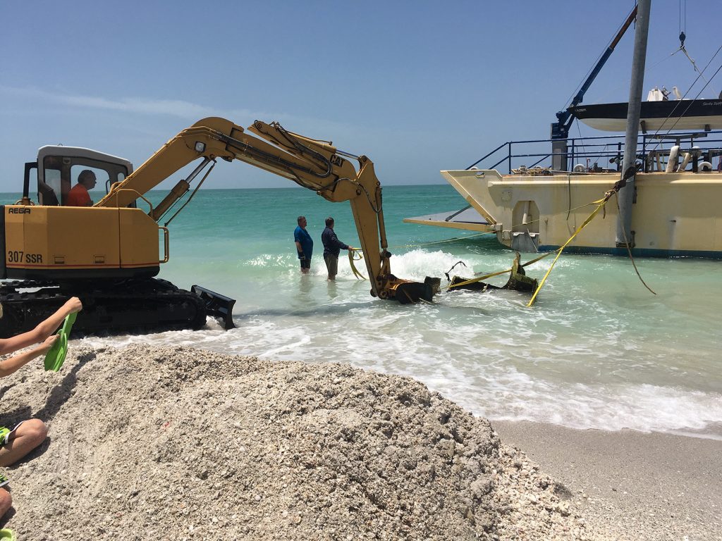 large backhoe digs into the sand and water on the beach. A large pile of sand sits on the shoreline