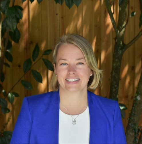 A photo of a blond woman wearing a blue jacket sitting in front of a fence smiling.