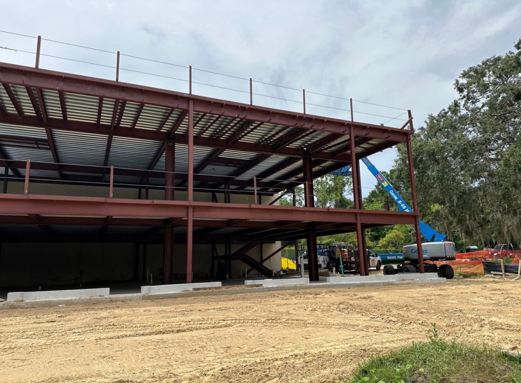 a construction area shows the large metal framing for a two story rectangular building with a lift extended tot he simple metal roof