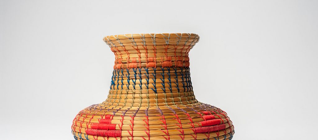 Basket with wide base and narrower neck woven with blue, red, green, orange and light blue thread. Simple designs woven from the blue, red, and orange thread decorate the basket.
