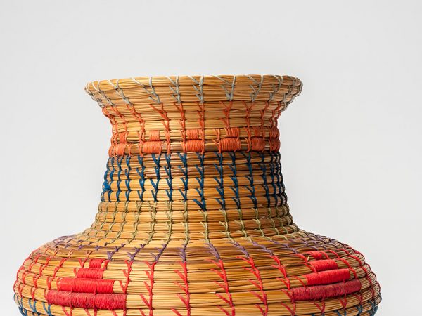 Basket with wide base and narrower neck woven with blue, red, green, orange and light blue thread. Simple designs woven from the blue, red, and orange thread decorate the basket.