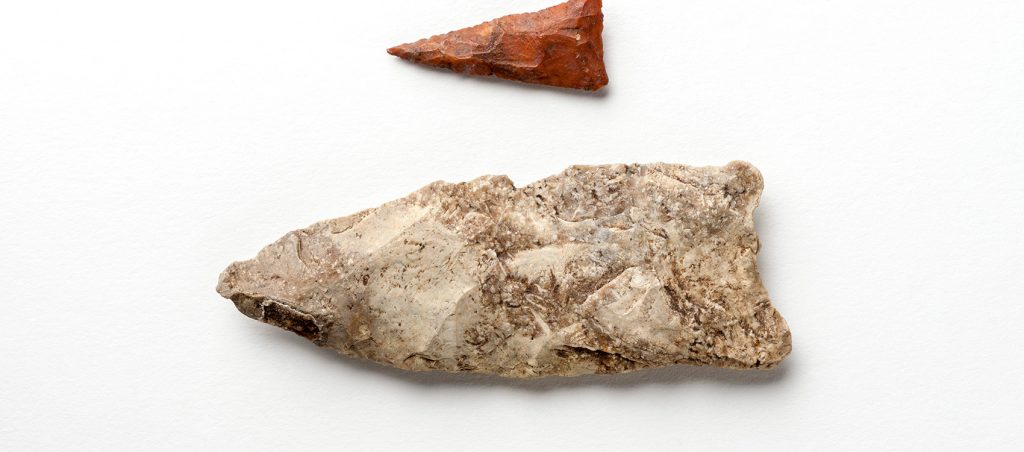 two Projectile Points showing the size difference. One is small red triangle with a long sharp point, the other is many times larger, one end squared off the other end sharpened to a point