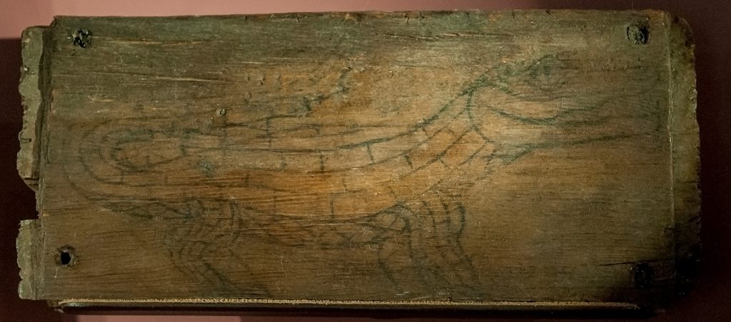 Flat piece of wood displayed and lit to show a faint line drawing of an alligator