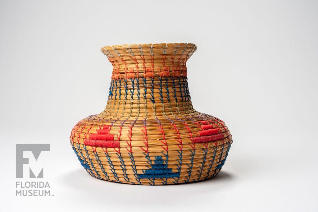 basket with wide base and narrower neck woven with blue, red, green, orange and light blue thread. Simple designs in blue, red, and orange decorate the basket.