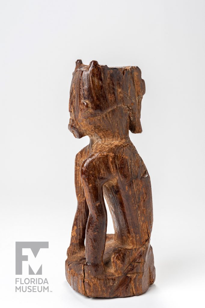 Carved wooden figurine with a squarish head, with well-defined facial features, torso and arms photographed from the side
