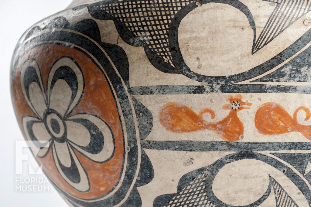 close up of vase painted with tan, black and Terra-cotta colored flowers, birds, and geometric patterns.