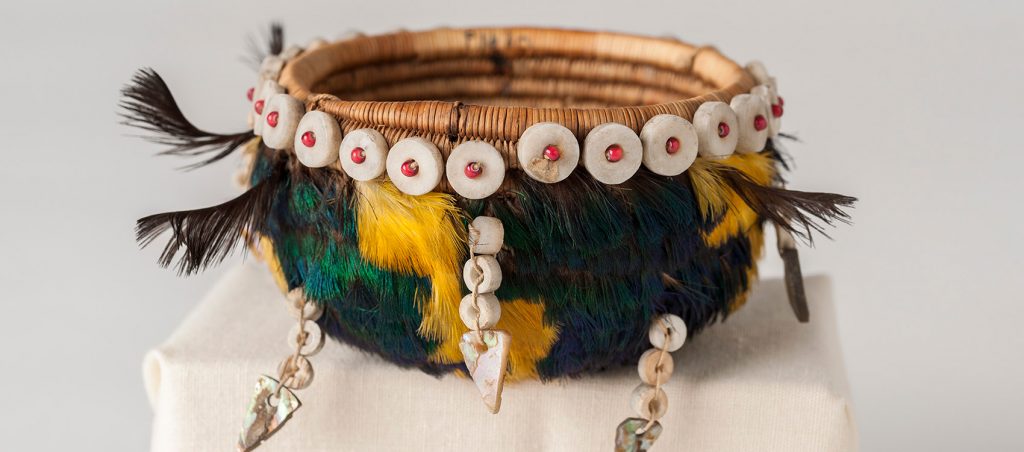 coiled basket decorated with bright yellow and blue-green feathers, shells and beads.