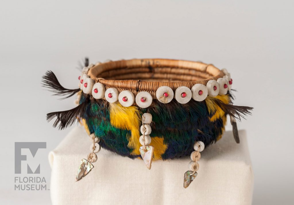 coiled basket decorated with bright yellow and blue-green feathers, shells, and beads.