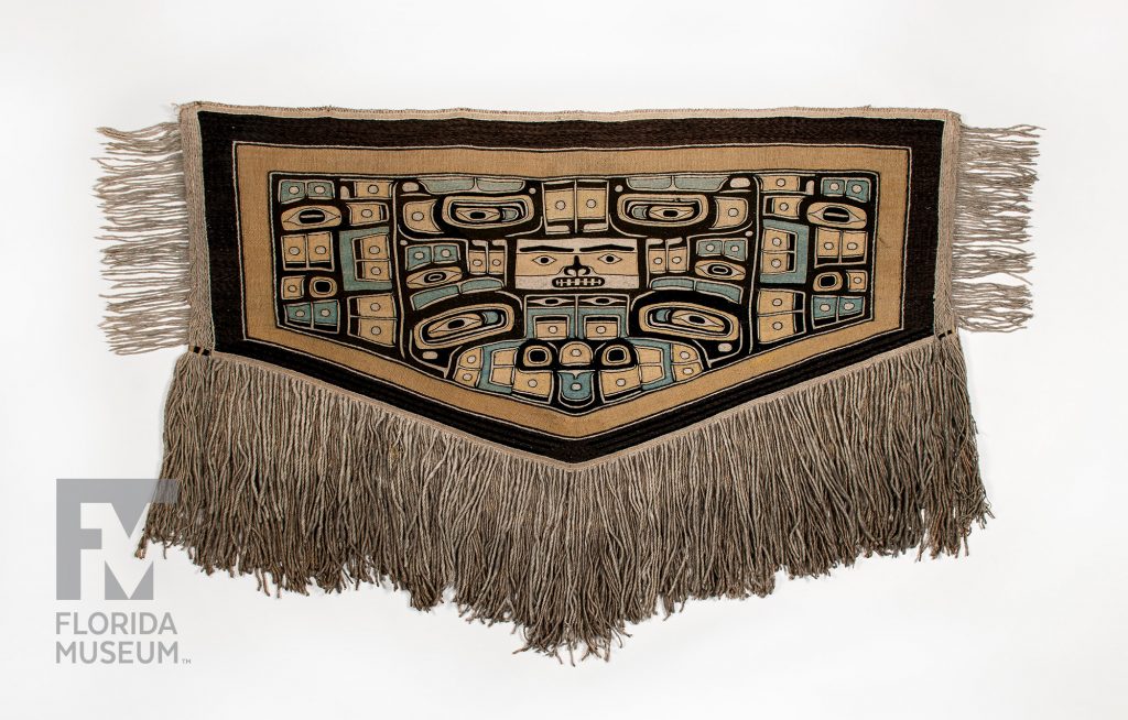woven blanket with motifs in tan, black, and blue. Long fringe hands from three edges of the blanket