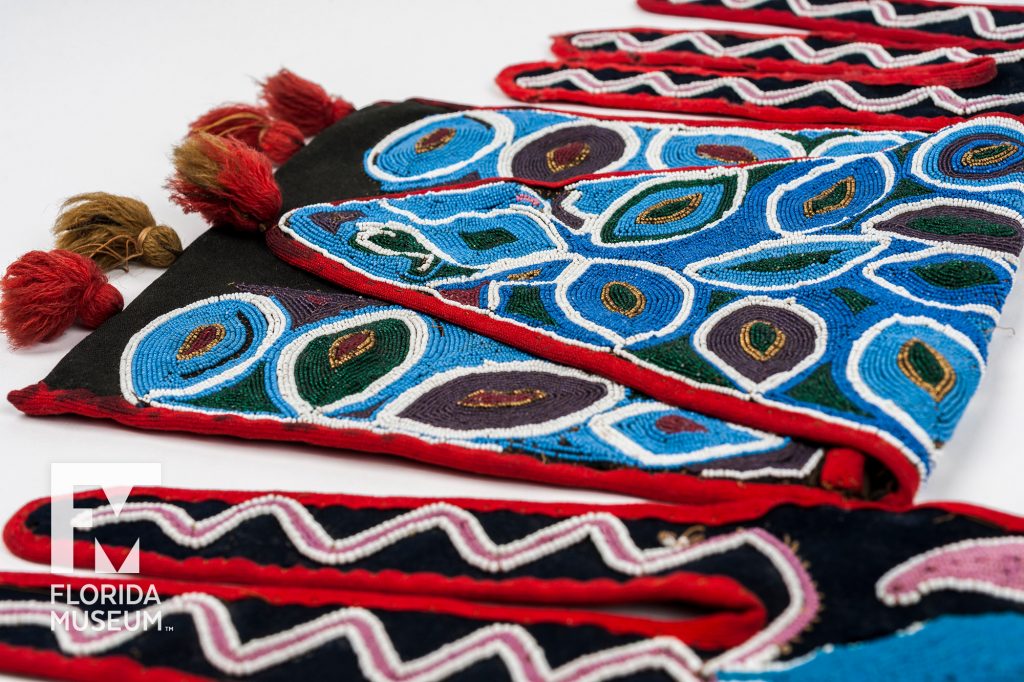 close up of brightly colored designs sewn from seed beads in shades of blue, green and maroon with gold accents. The edges of the fabric are red and tassels decorated to bottom of the bag