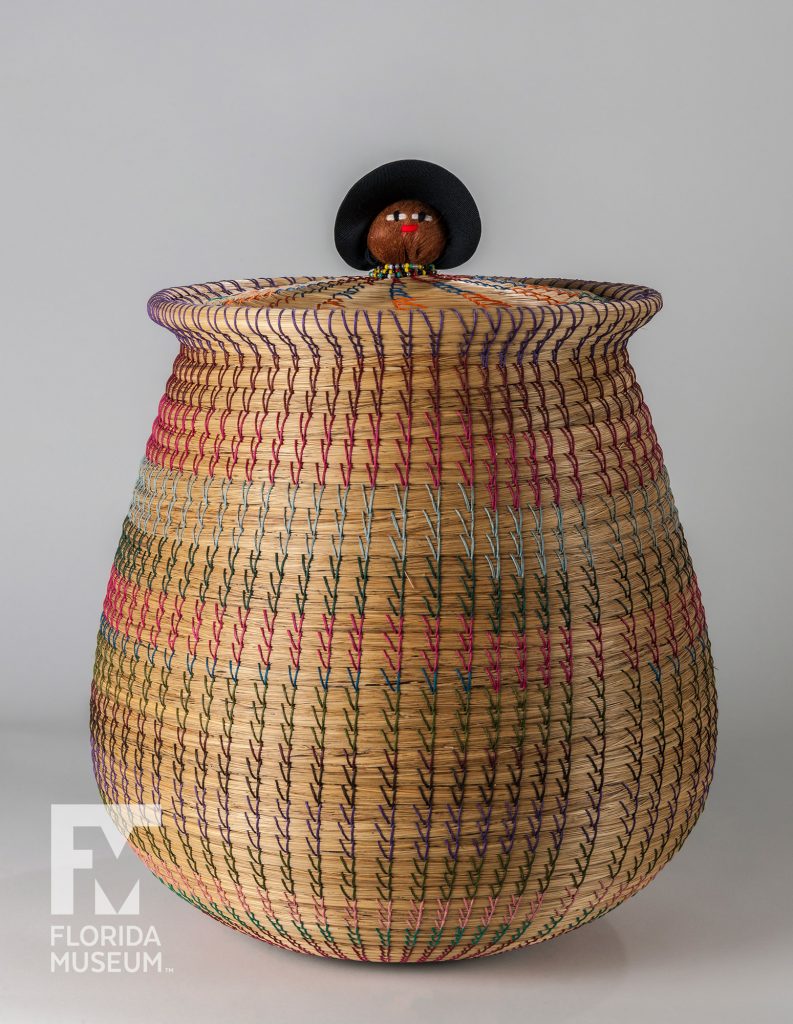 coiled woven basket made from tan sweet grass and multi colored cotton thread. At the center of the basket's lid is a doll head.