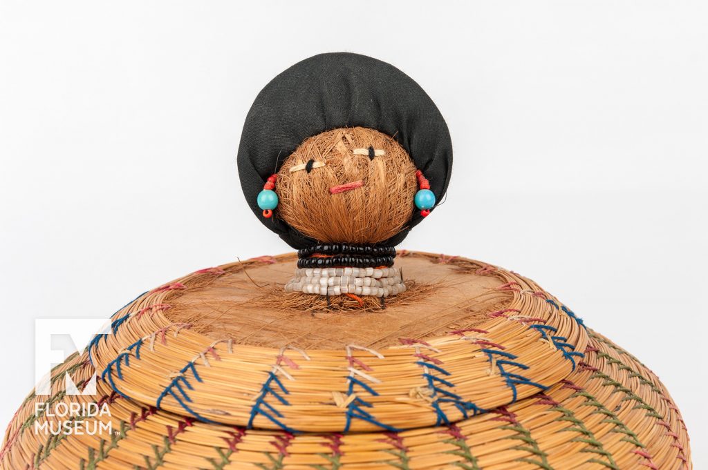 close up of the basket lid. At the center of the lid is a doll head with a beaded face and jewelry