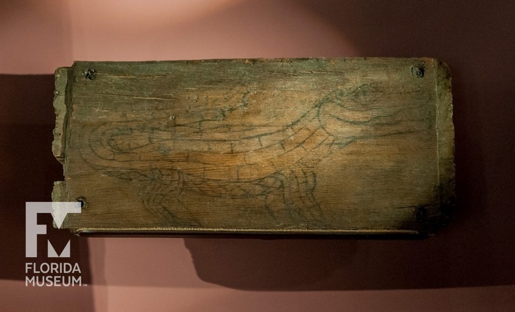 Flat piece of wood displayed and lit to show a faint line drawing of an alligator