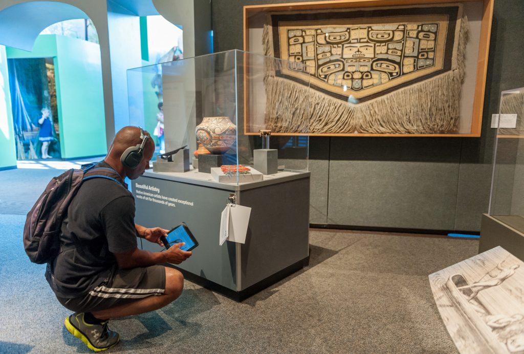 A visitor kneels next it an item on display while holding a tablet and listening to headphones each item on display includes an audio narration.