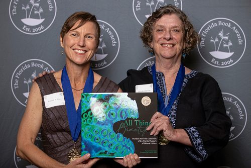 two people stand facing the camera and smiling. They each wear award metals and are holding the book All Things Beautiful