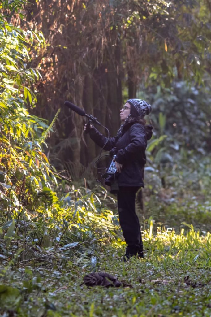 Glaucia Del-Rio holding a large microphone and recording equipment standing in the woods