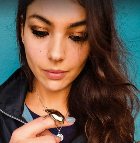 An image of a woman, Chelsea Skojec, holding a golden beetle. The background of the image is blue.
