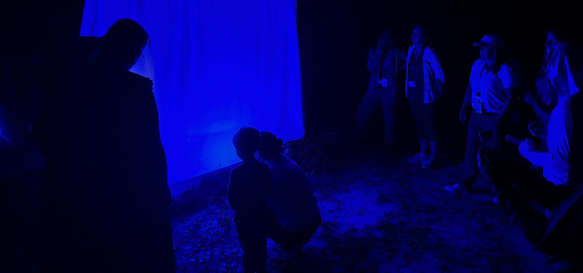 Multiple people gathered in front of a light sheet with a black light casting a deep blue hue.