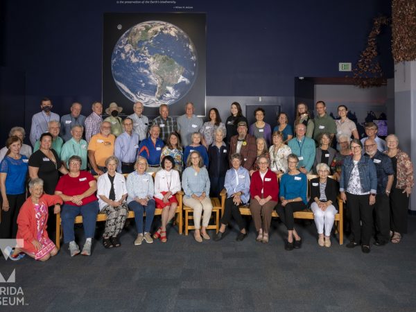 Around 45 volunteers gather in 3 rows in the Butterfly Rainforest gallery with a large map of earth in the background.