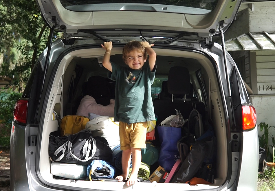 child standing in the back of a car with packed camping equipment behind them