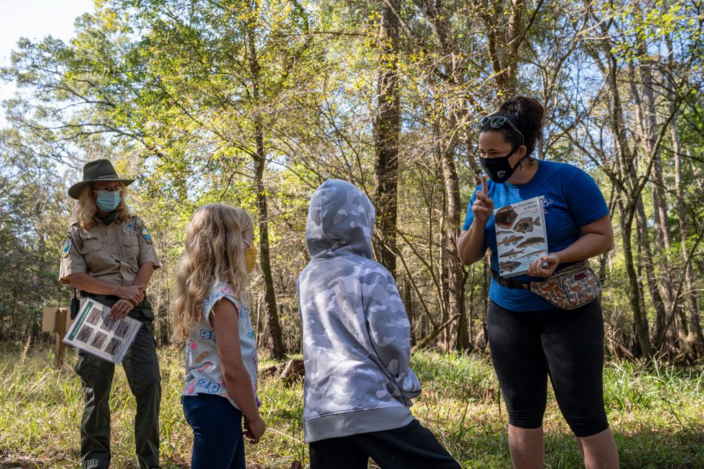 tour guild hold book Fishes of the Fresh Waters of Florida and speaks to two children, in the background a park ranger watches