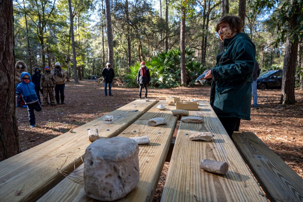 masked tour guild stands by a picnic table with display items, rocks and fossils, spread out. She is speaking to a group of people standing socially distanced around the park