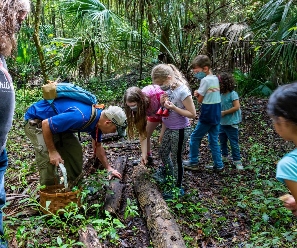 several children and a researcher look under a log for mushrooms