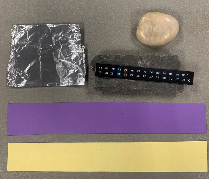 collection of objects for reptile basking activity: piece of Aluminum foil, two rocks, one tan and one grey, two pieces of paper, one purple one yellow, Flat sticker thermometer