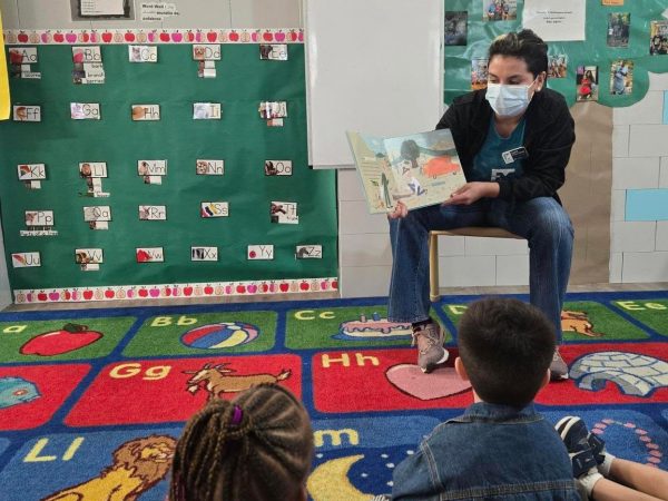 Education team member sits on a chair and holds a book she is reading to a group of children
