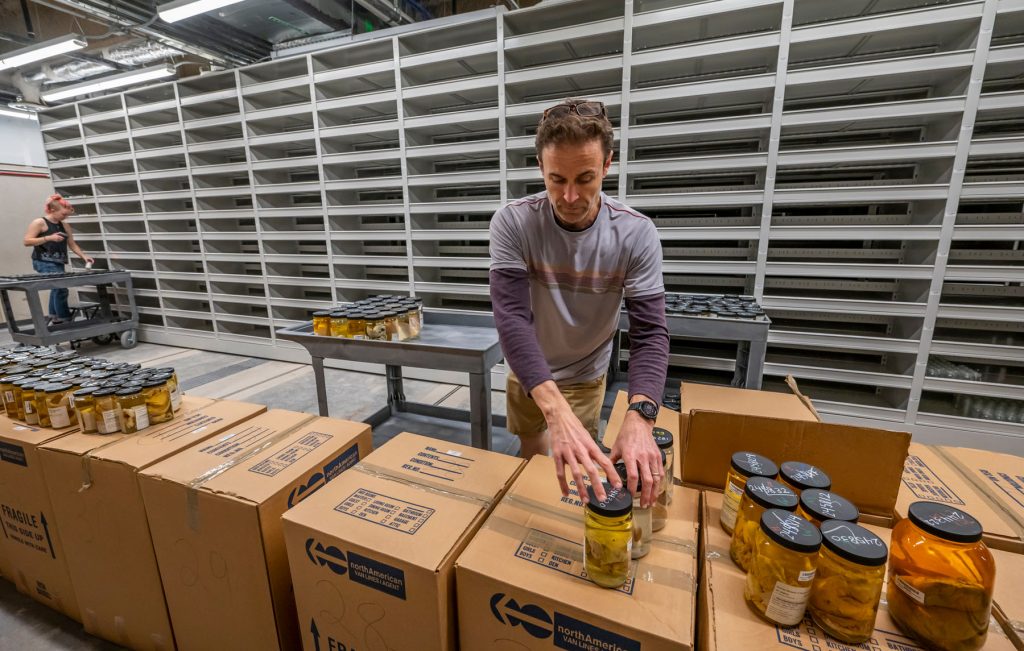 A man sorts glass jars filled with scientific specimens, moving them from on top of stacked cardboard boxes to a cart. Behind him are rows of tall empty shelves. In the background another person moves more glass jars.