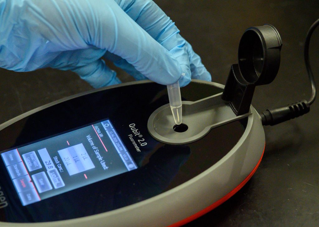 a hand in a blue medical glove pushes a small plastic vial into the hole of a oval device with a digital readout
