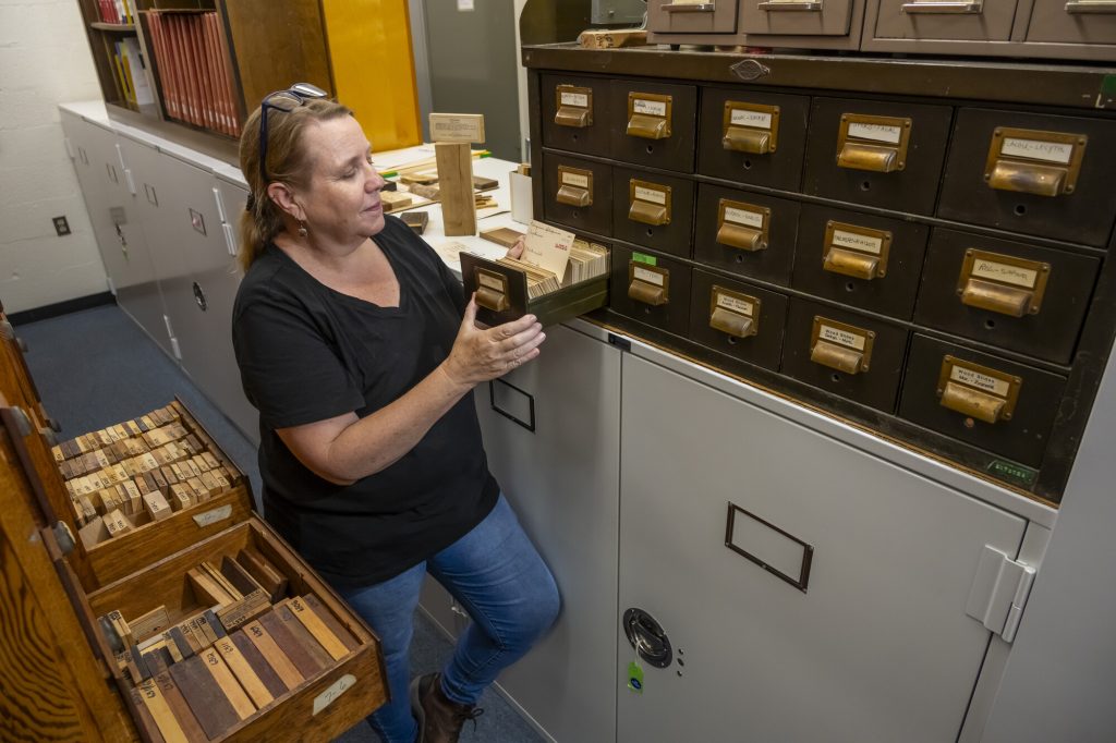 a person is standing on a narrow library corridor looking through a card catalog on one side and holing open drawers full of short planks of wood