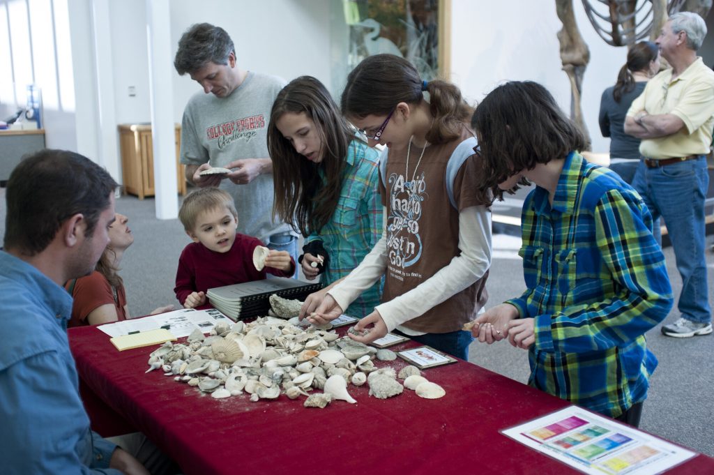 a group of children are gathered around a table in a museum to look at a pile of shells and talk to several adults about them
