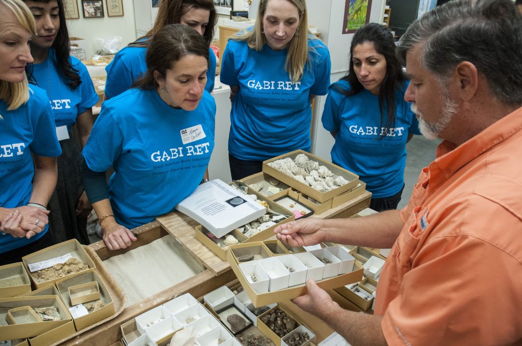 a group of people in matching t-shirts are standing at a table while a man holds up one of several trays of shallow boxes with different types of shells sorted neatly into them