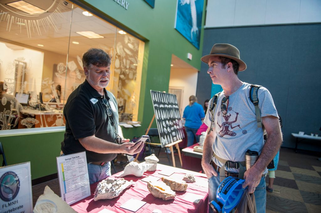 two people are standing over a table covered in fossils in a sunny open space in a museum lobby