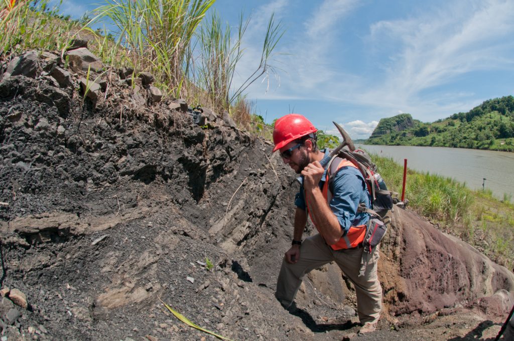 man in helmet swings pickaxe at dirt with canal in background